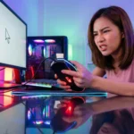 หญิงสาวเกมเมอร์ทำหน้าหงุดหงิดโมโหขณะถือเมาส์ที่มีไฟแต่ขยับไม่ได้ หน้าจอคอมพิวเตอร์แสดงผลค้าง ในห้องไฟ RGB
