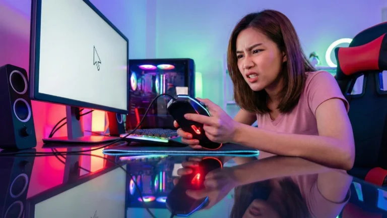 หญิงสาวเกมเมอร์ทำหน้าหงุดหงิดโมโหขณะถือเมาส์ที่มีไฟแต่ขยับไม่ได้ หน้าจอคอมพิวเตอร์แสดงผลค้าง ในห้องไฟ RGB